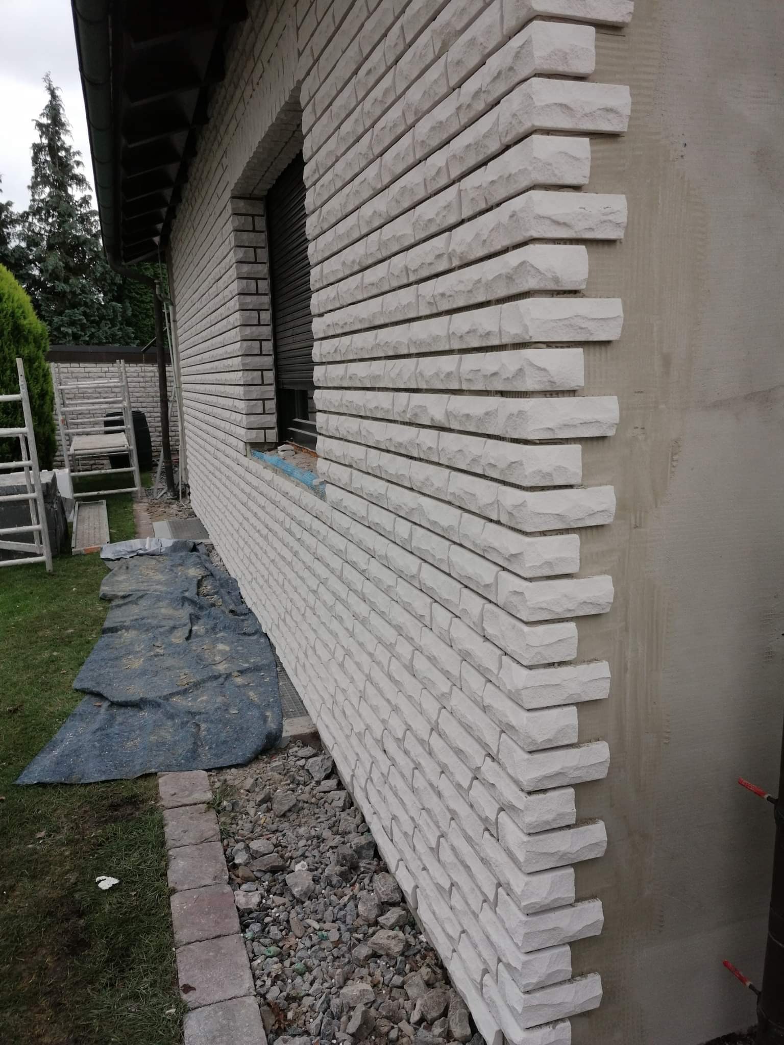 Ściana budynku z częściowo ułożoną elewacją z białego kamienia, widoczne niedokończone tynkowanie narożnika i rozłożona folia ochronna na trawniku.