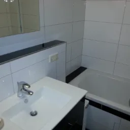 Biała łazienka z wanną i umywalką, wykończona białymi płytkami z czarnymi akcentami, widoczny fragment lustra i mydelniczka z szarym mydłem