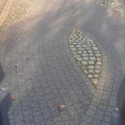 Zakrzywiony chodnik z kostki brukowej w parku, z dekoracyjnym wzorem w kształcie liścia wykonanym z innej kostki, częściowo przysłonięty przez fragment ogrodzenia.