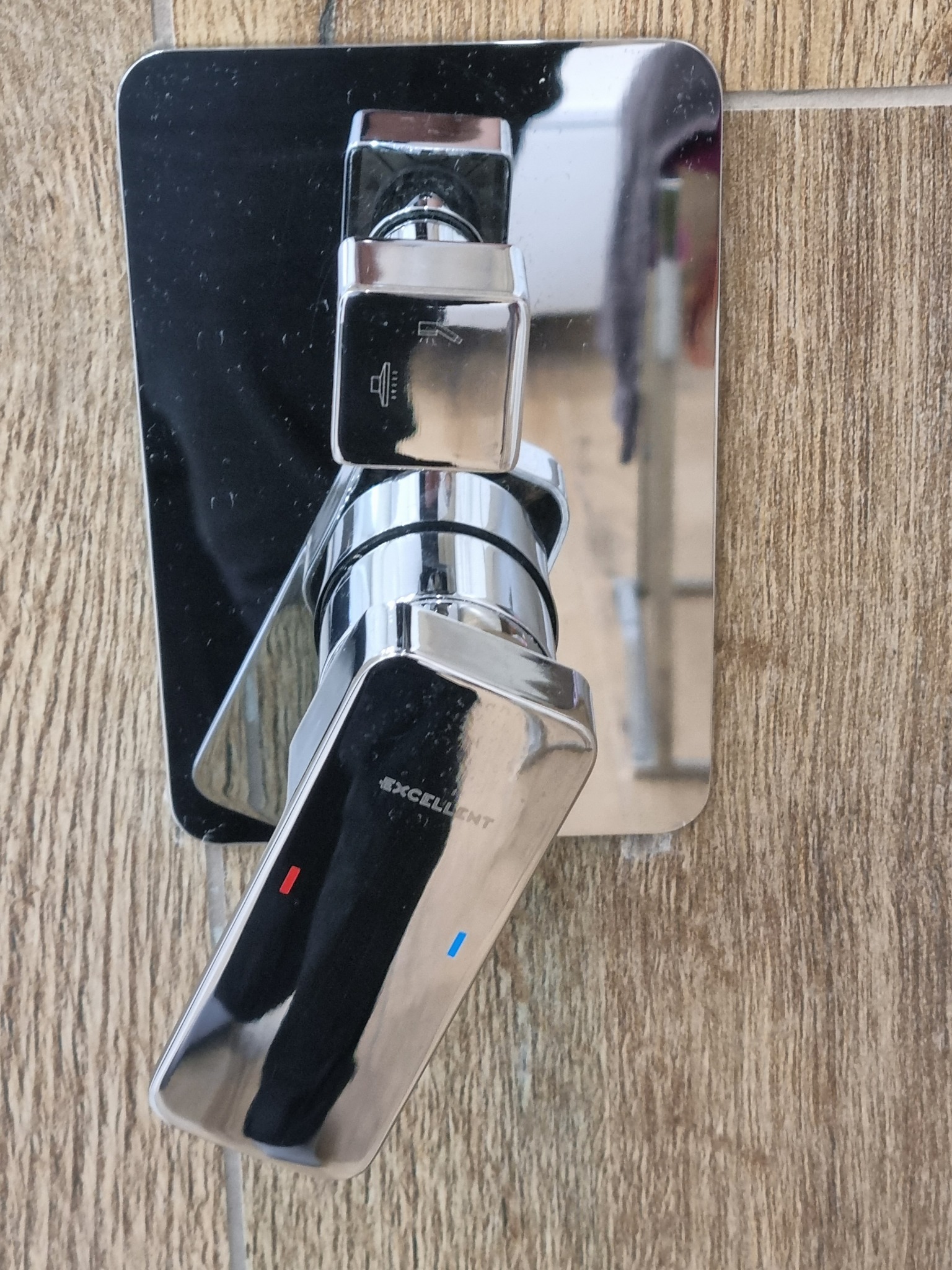 Błyszcząca, chromowana bateria natryskowa marki Excellent zamontowana na ścianie wyłożonej płytkami imitującymi drewno.