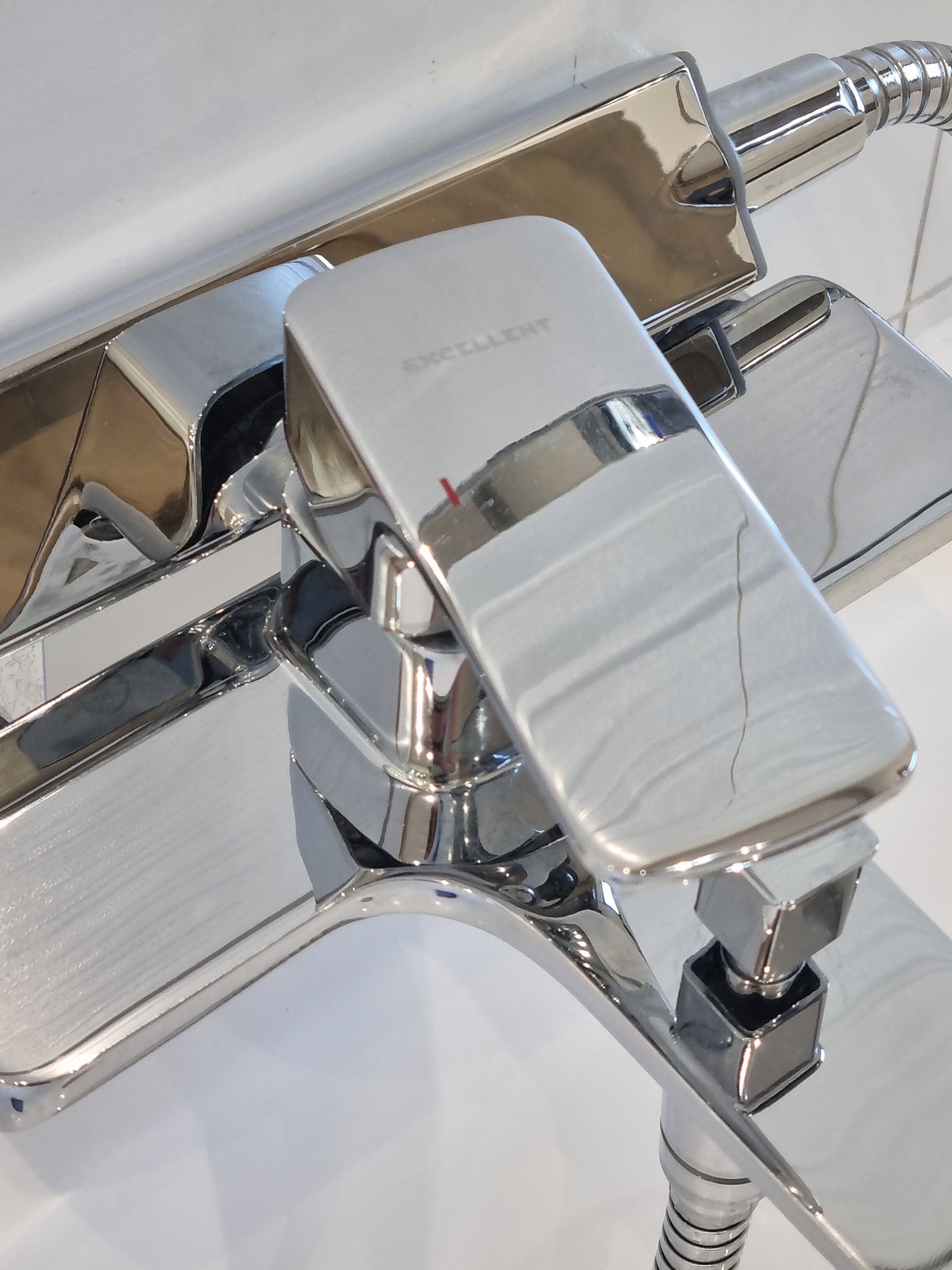 Błyszcząca, chromowana bateria wannowa marki Excellent zamontowana na białej wannie, widoczny wąż prysznicowy i odbicia światła na powierzchni armatury.