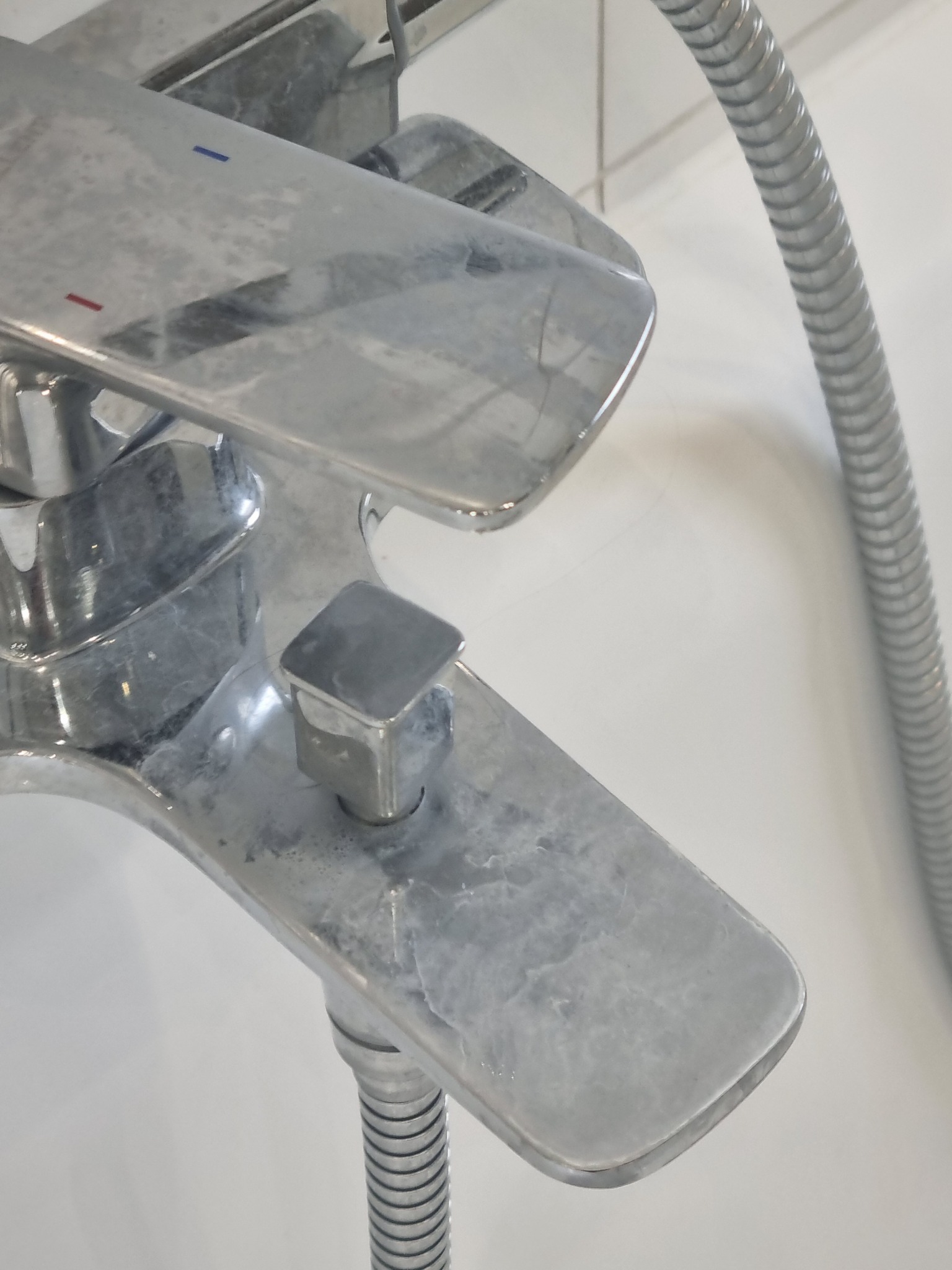 Chromowana bateria wannowa z widocznymi śladami osadu wapiennego na powierzchni, obok elastyczny wąż prysznicowy.