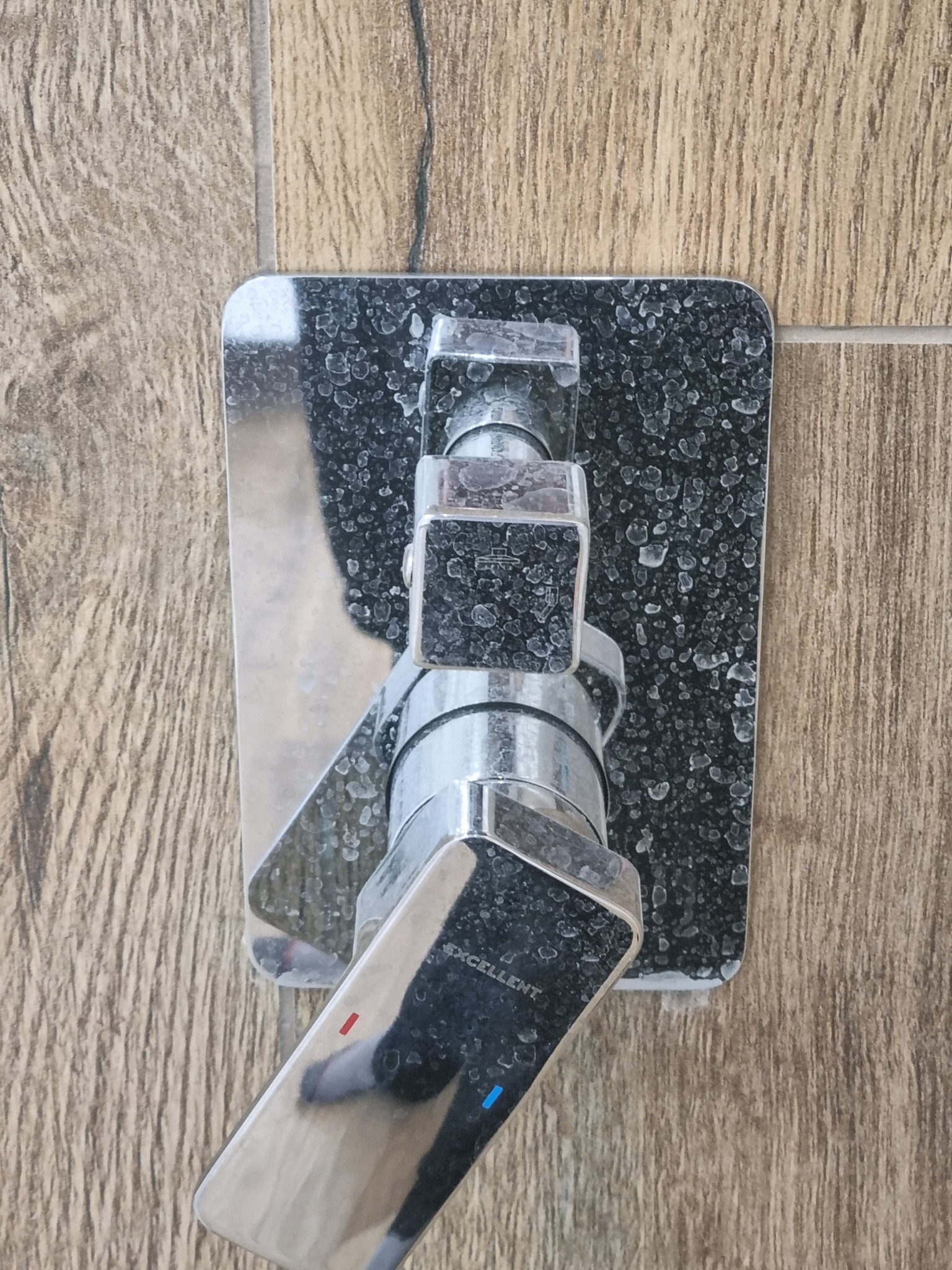 Bateria umywalkowa chromowana, pokryta osadem z kamienia, zamontowana na ścianie wyłożonej płytkami imitującymi drewno.