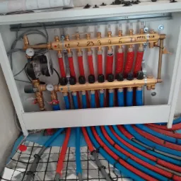 Rozdzielacz ogrzewania podłogowego TECE w szafce, z widocznymi rurami w kolorach czerwonym i niebieskim, ułożonymi na izolacji podłogowej z folii aluminiowej w kratkę, z planem instalacji na górnej...