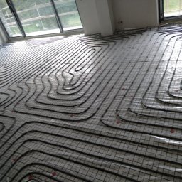 NOVO-TECH Sławomir Czmiel - Rozłożone rury ogrzewania podłogowego na siatce montażowej w jasnym pomieszczeniu z dużymi oknami, widoczne czerwone elementy mocujące.