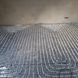 NOVO-TECH Sławomir Czmiel - Rozłożona instalacja ogrzewania podłogowego z białych rurek na srebrnej folii z czarną siatką, zabezpieczonych czarnymi opaskami zaciskowymi, widoczne trzy puszki elektryczne w suficie.