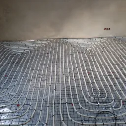 Rozłożona instalacja ogrzewania podłogowego z białych rurek na srebrnej folii z czarną siatką, zabezpieczonych czarnymi opaskami zaciskowymi, widoczne trzy puszki elektryczne w suficie.