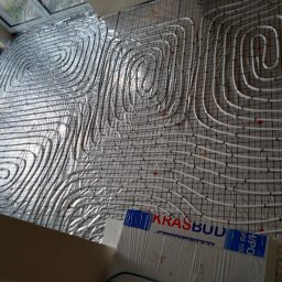 NOVO-TECH Sławomir Czmiel - Rozłożona instalacja ogrzewania podłogowego z białych rurek na metalowej siatce, widok z góry, fragment okna i izolacji z logiem firmy budowlanej w dolnej części kadru.