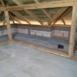 NOVO-TECH Sławomir Czmiel - Szare rury karbowane ułożone równolegle na betonowej posadzce w surowym stanie, widoczne drewniane elementy konstrukcyjne dachu i ściany z bloczków betonowych.