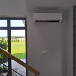 NOVO-TECH Sławomir Czmiel - Biały klimatyzator LG Dual Inverter zamontowany na szarej ścianie w nowoczesnym wnętrzu, widok z klatki schodowej na ogród przez duże okno.