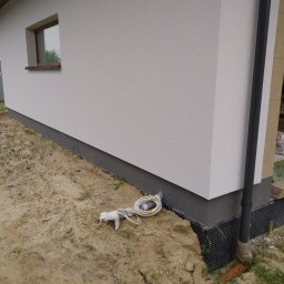 NOVO-TECH Sławomir Czmiel - Fragment budynku z oknem i rurą spustową. Na ziemi leżą zwinięte kable i rura owinięta taśmą. Widoczna izolacja fundamentów z folii kubełkowej.