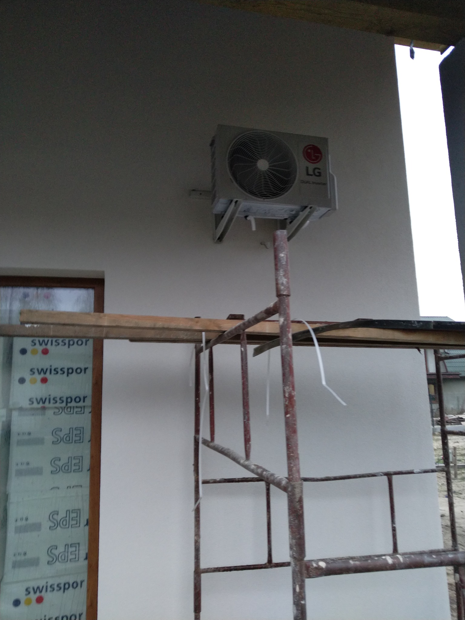 Zewnętrzna jednostka klimatyzacji LG Dual Inverter zamontowana na elewacji budynku w Woli Paprotni, widoczna z poziomu rusztowania. Obok okno z opakowaniami styropianu Swissopor.