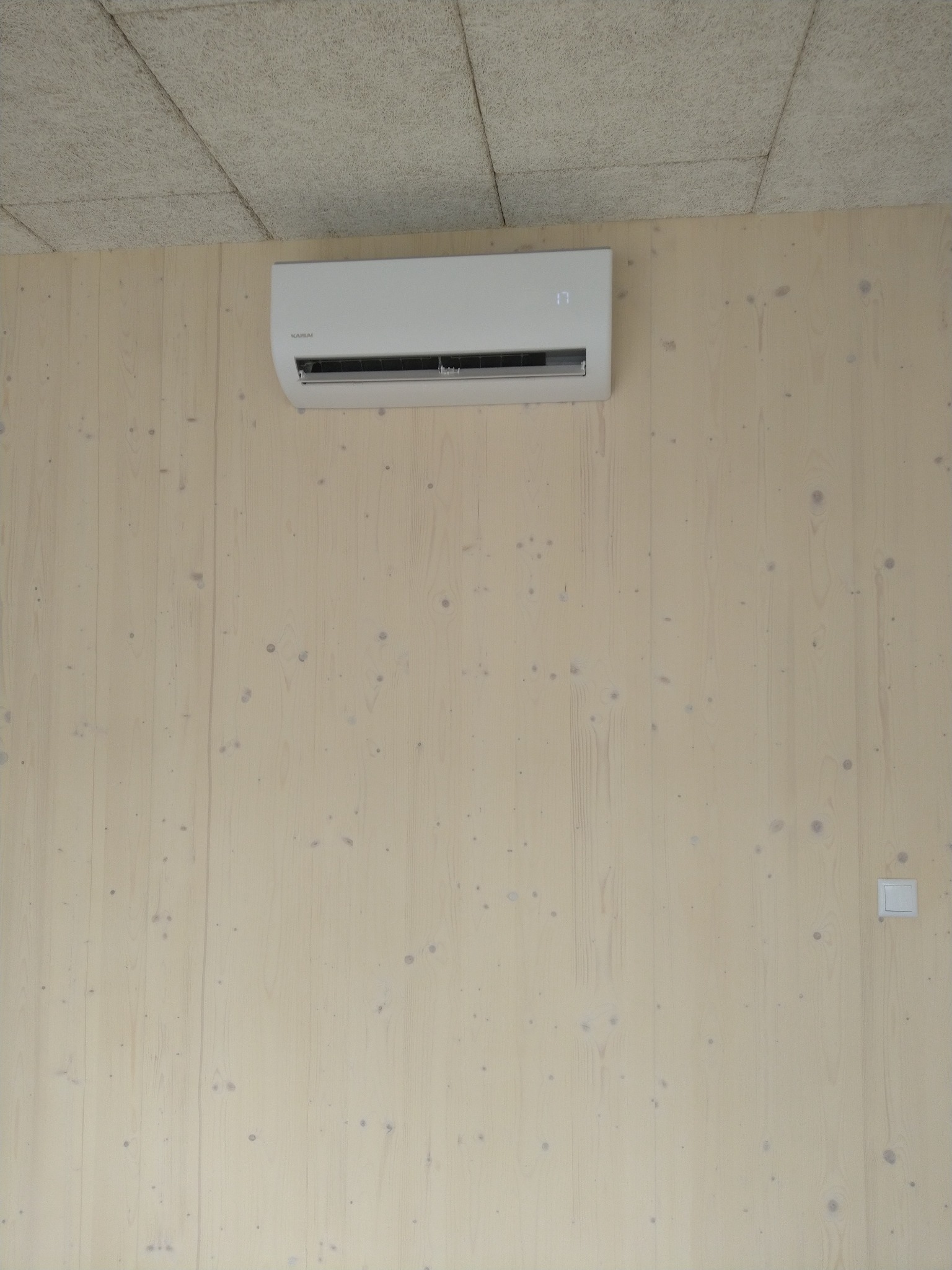 Biała klimatyzacja typu split zamontowana na jasnej, drewnianej ścianie z widocznym wyświetlaczem temperatury i wyłącznikiem światła w prawym dolnym rogu.