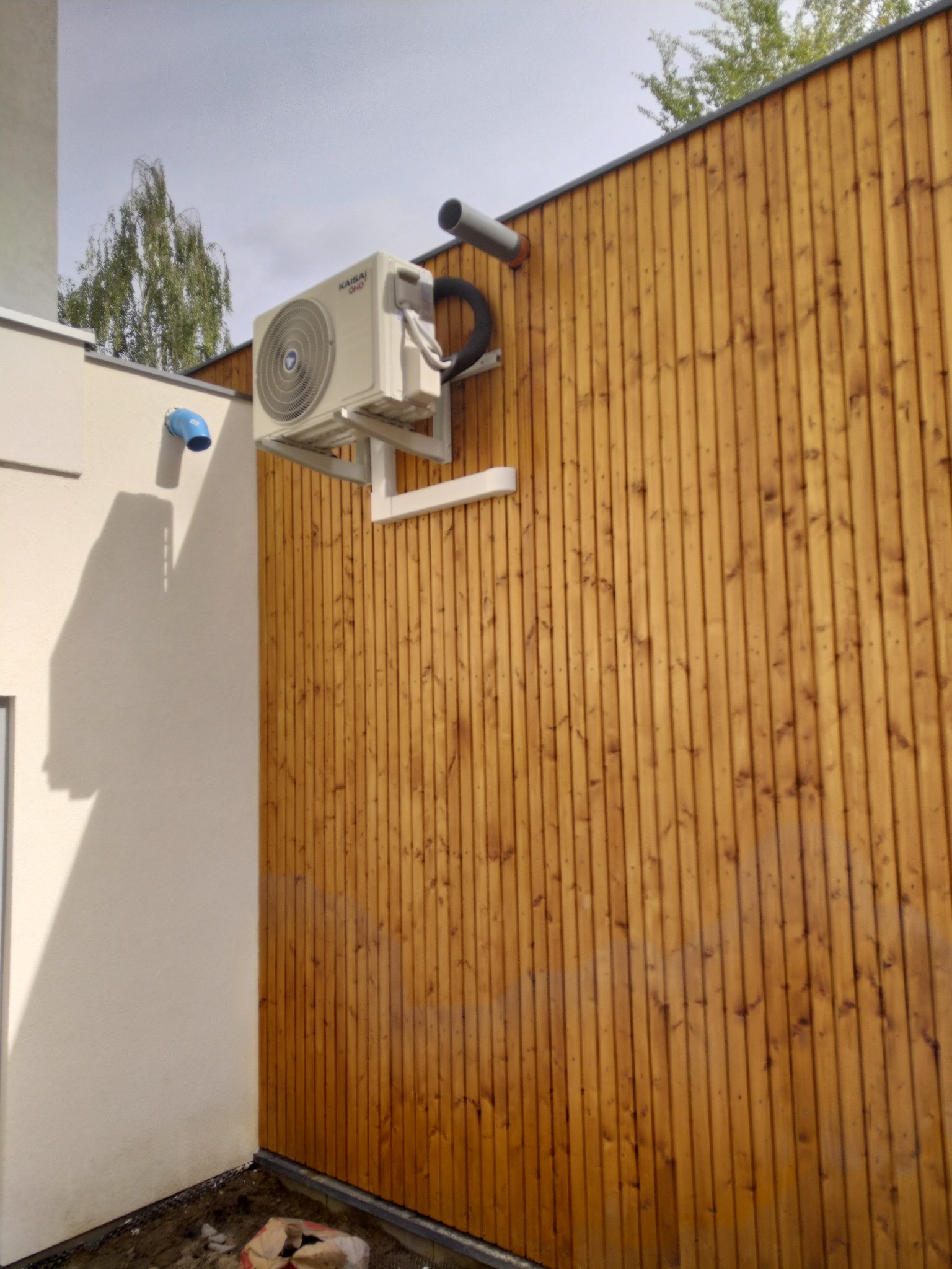 Zewnętrzna jednostka klimatyzacji marki Kaisai zamontowana na białym wsporniku na drewnianej ścianie z pionowych desek, widoczny fragment rury odprowadzającej skropliny oraz element instalacji...