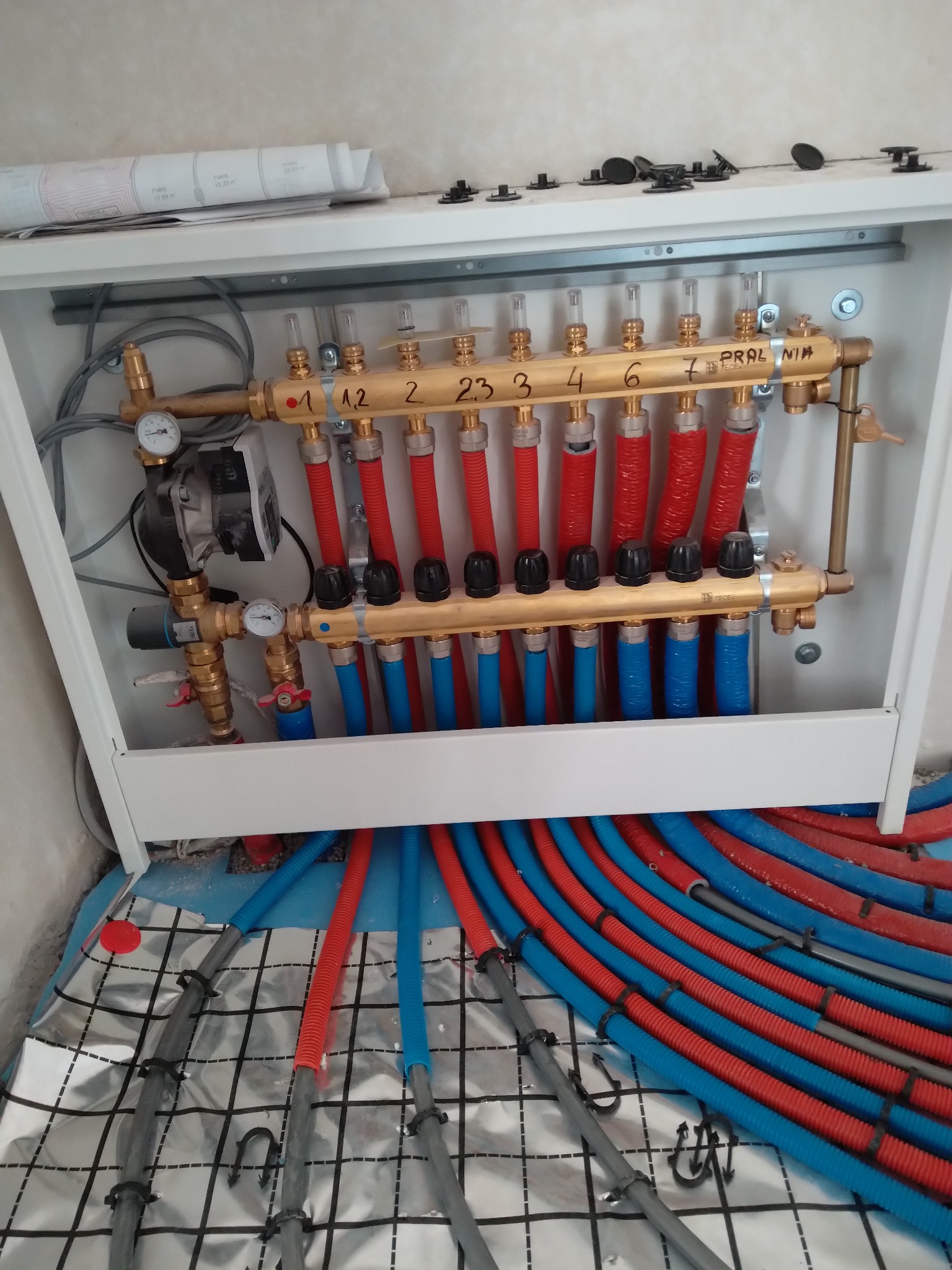 Rozdzielacz ogrzewania podłogowego TECE w szafce, z widocznymi rurami w kolorach czerwonym i niebieskim, ułożonymi na izolacji podłogowej z folii aluminiowej w kratkę, z planem instalacji na górnej...