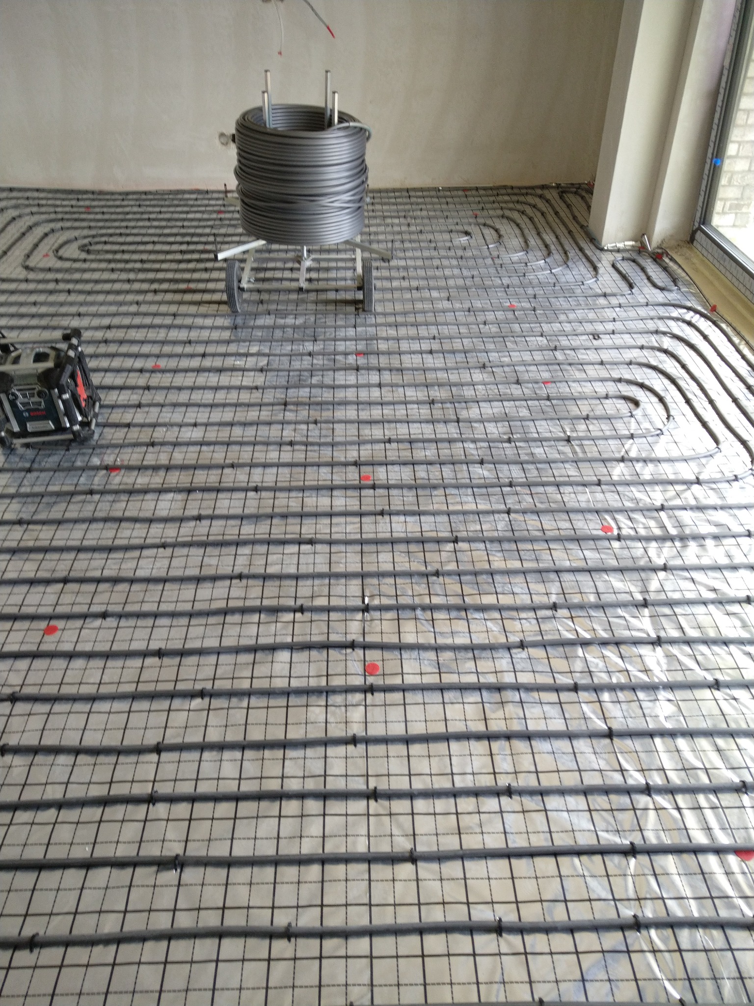 Instalacja ogrzewania podłogowego: rury grzewcze ułożone na folii izolacyjnej, przymocowane do siatki, z rolką rur na wózku w tle i poziomnicą laserową po lewej stronie.