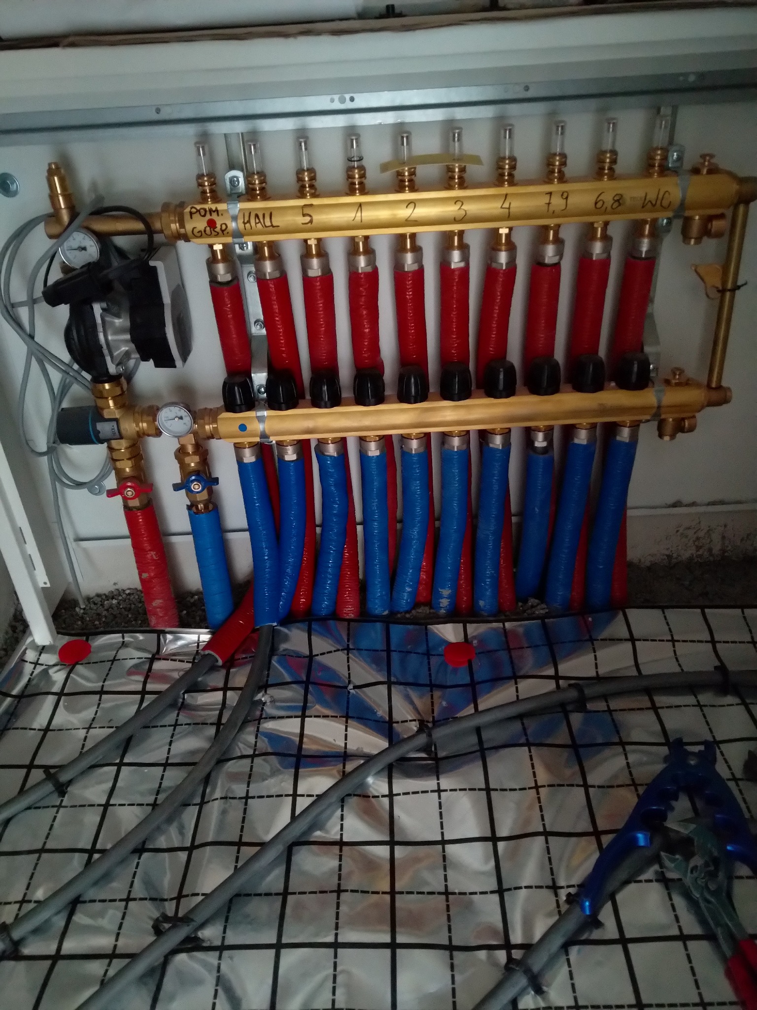 Rozdzielacz podłogowy z rurami w kolorach czerwonym i niebieskim, oznaczony nazwami pomieszczeń, zamontowany w szafce, z widocznymi narzędziami do montażu.