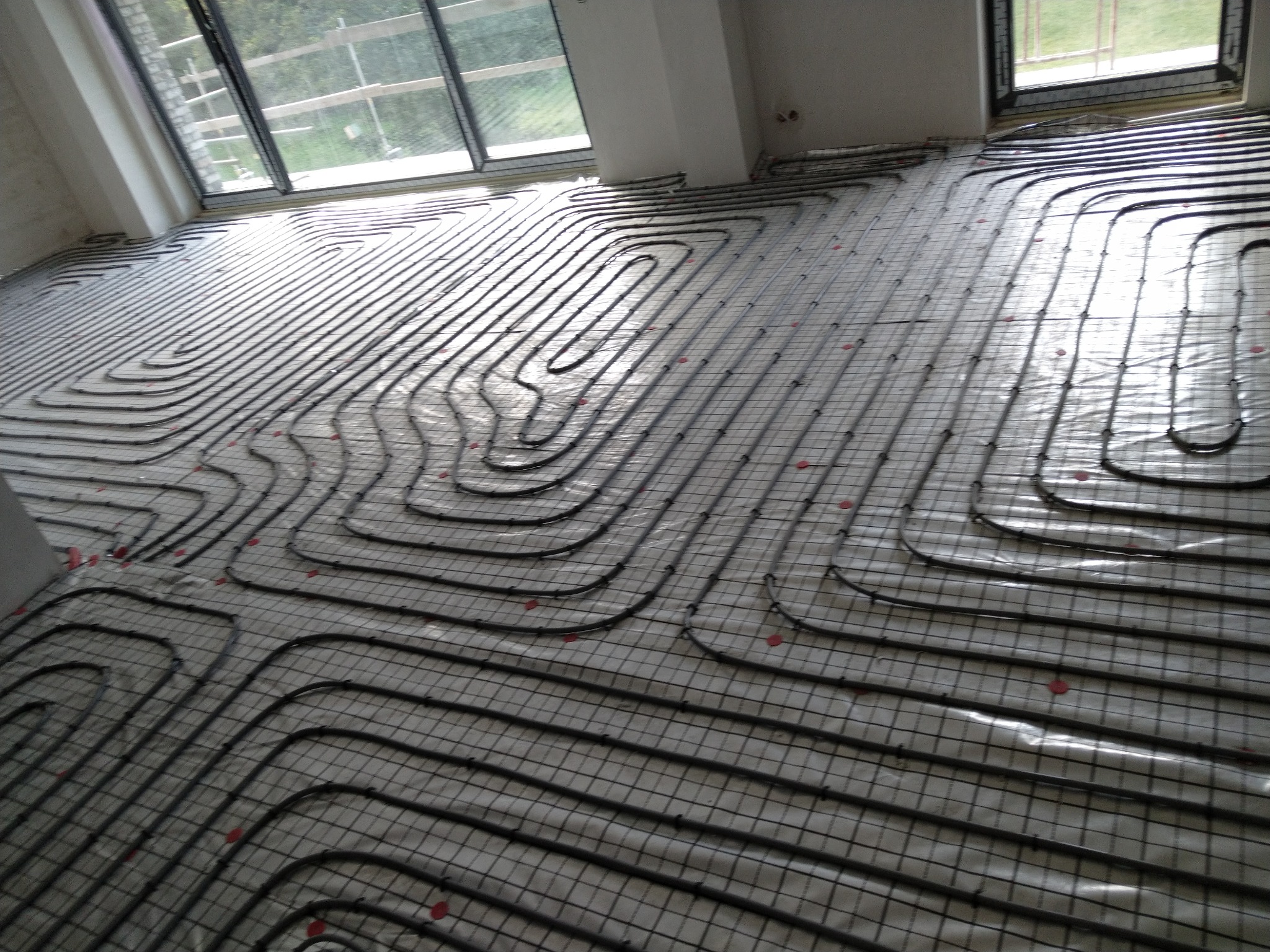 Rozłożone rury ogrzewania podłogowego na siatce montażowej w jasnym pomieszczeniu z dużymi oknami, widoczne czerwone elementy mocujące.