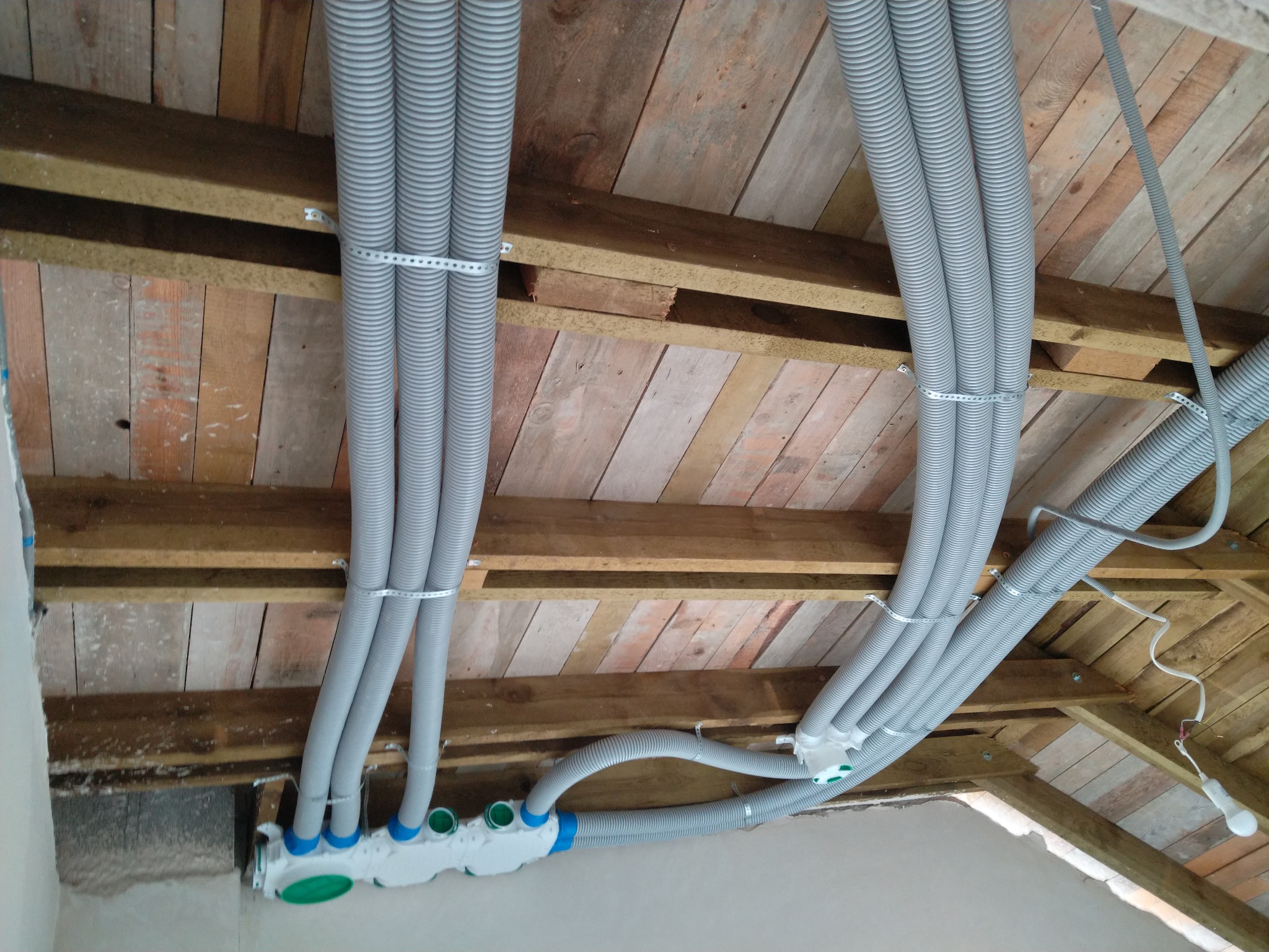 Szare, elastyczne przewody wentylacyjne zamontowane pod drewnianym stropem, mocowane metalowymi opaskami. Widoczny rozdzielacz z niebieskimi i zielonymi elementami.