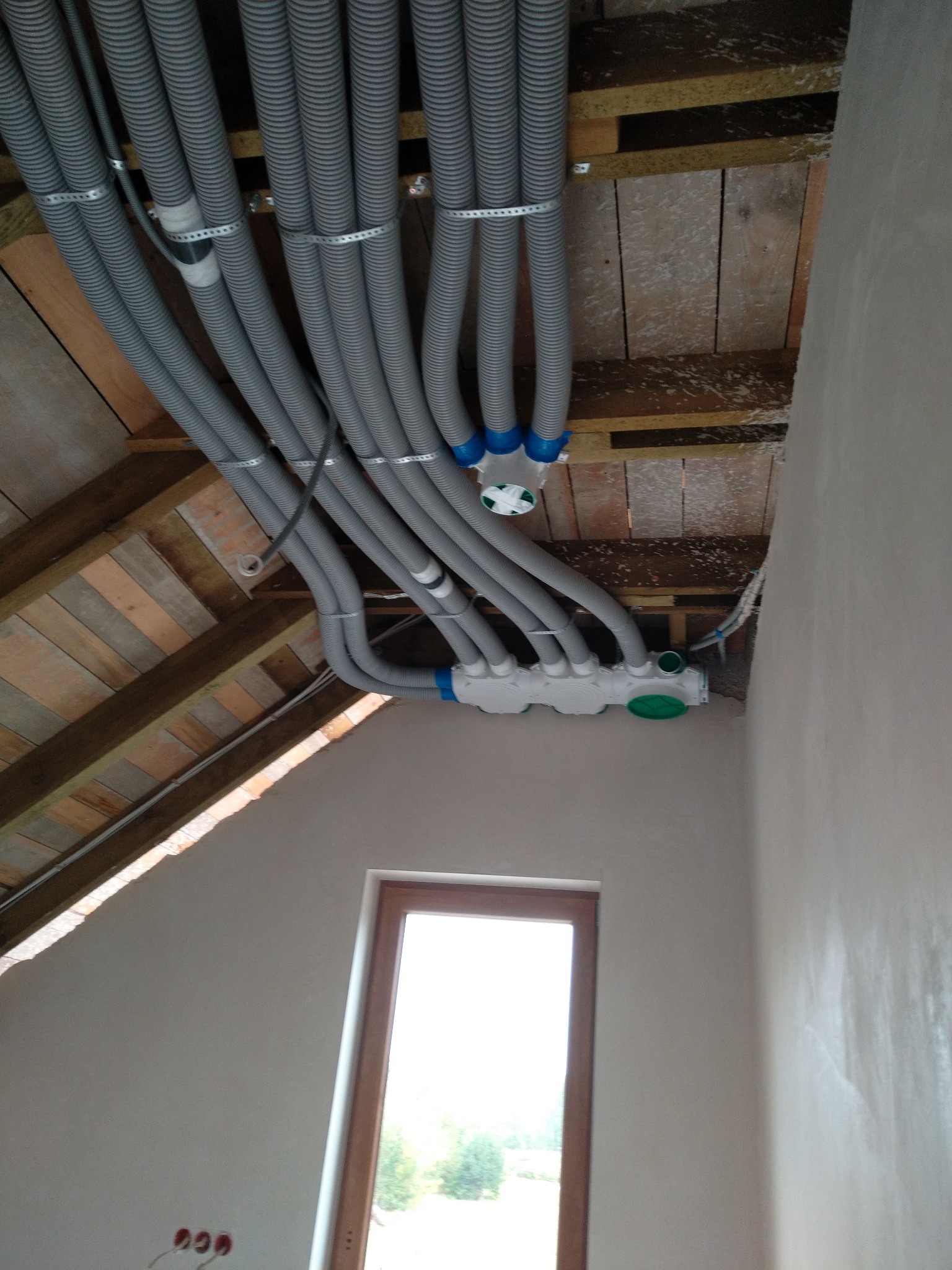 Szare, karbowane rury wentylacyjne przymocowane do drewnianej konstrukcji dachu na poddaszu, z widocznymi elementami łączeniowymi i oknem w tle.