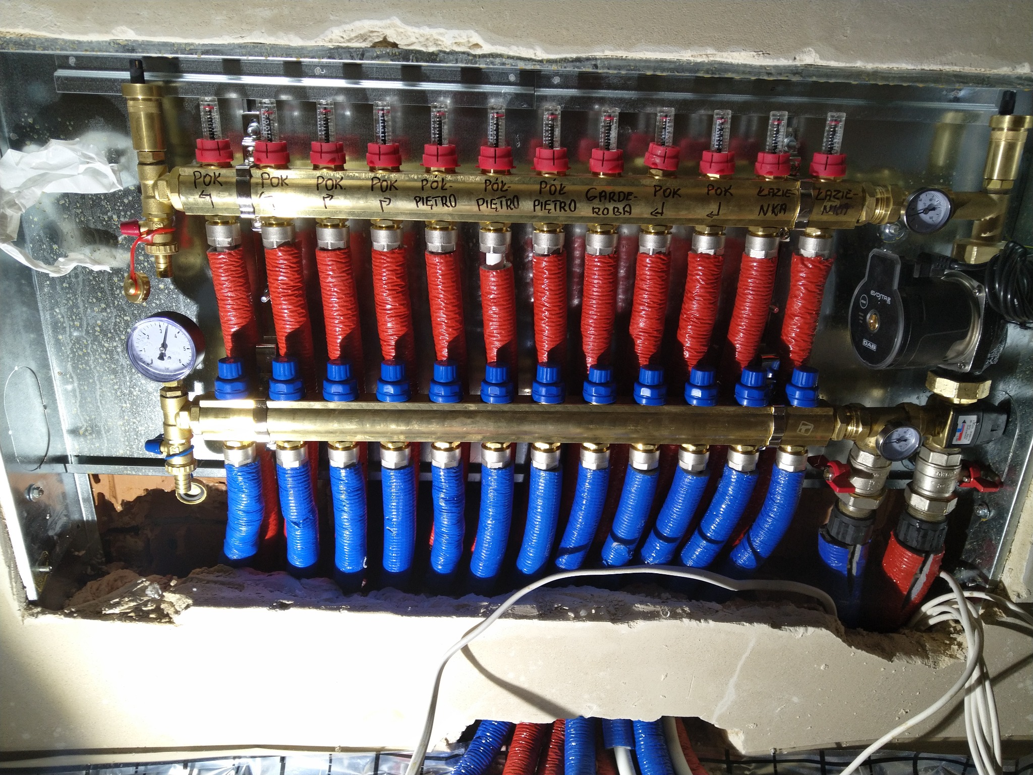 Rozdzielacz ogrzewania podłogowego z rurami w izolacji czerwonej i niebieskiej, zamontowany w obudowie wnęki ściennej, widoczne manometry i przepływomierze.