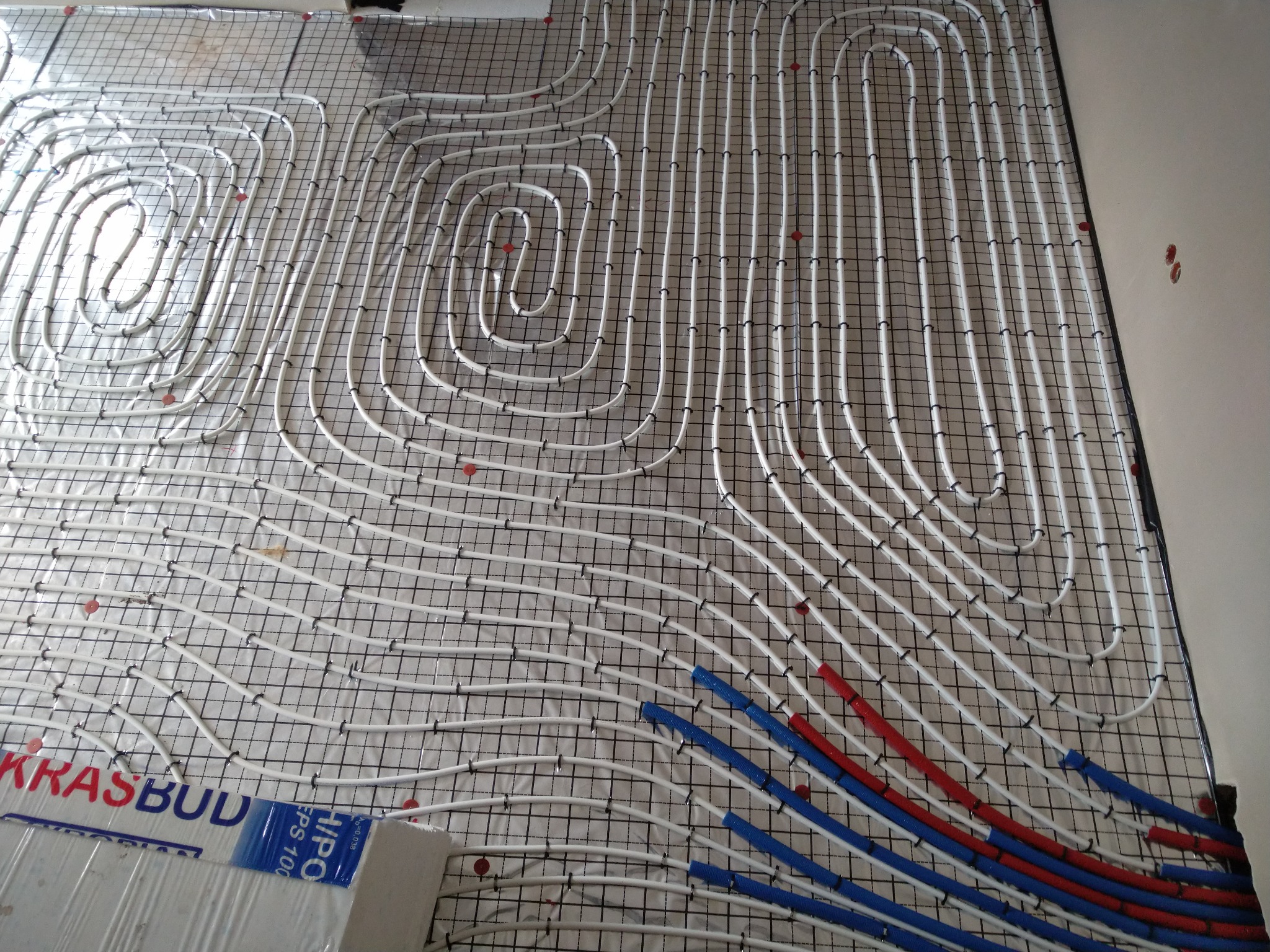 Rozłożone białe rury ogrzewania podłogowego na siatce z czarnych prętów, z fragmentami rur oznaczonych kolorem niebieskim i czerwonym, widoczne fragmenty styropianu z nadrukiem 'KRASBUD'.