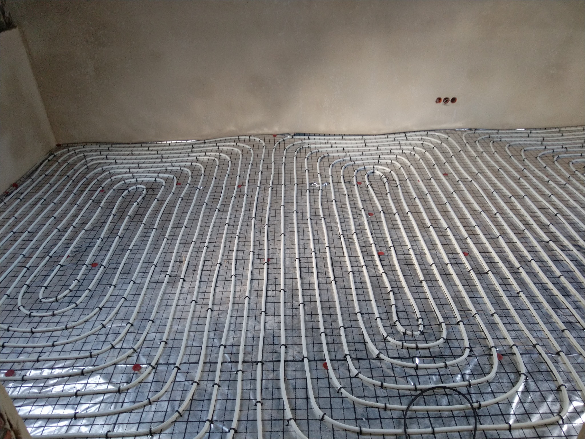 Rozłożona instalacja ogrzewania podłogowego z białych rurek na srebrnej folii z czarną siatką, zabezpieczonych czarnymi opaskami zaciskowymi, widoczne trzy puszki elektryczne w suficie.