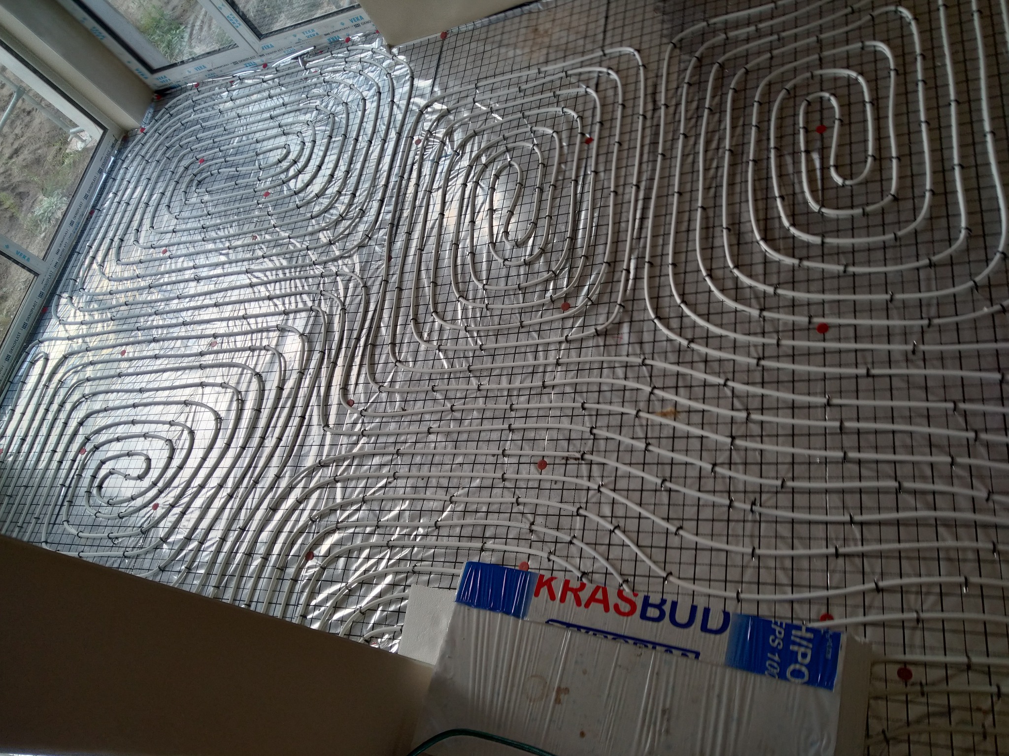 Rozłożona instalacja ogrzewania podłogowego z białych rurek na metalowej siatce, widok z góry, fragment okna i izolacji z logiem firmy budowlanej w dolnej części kadru.