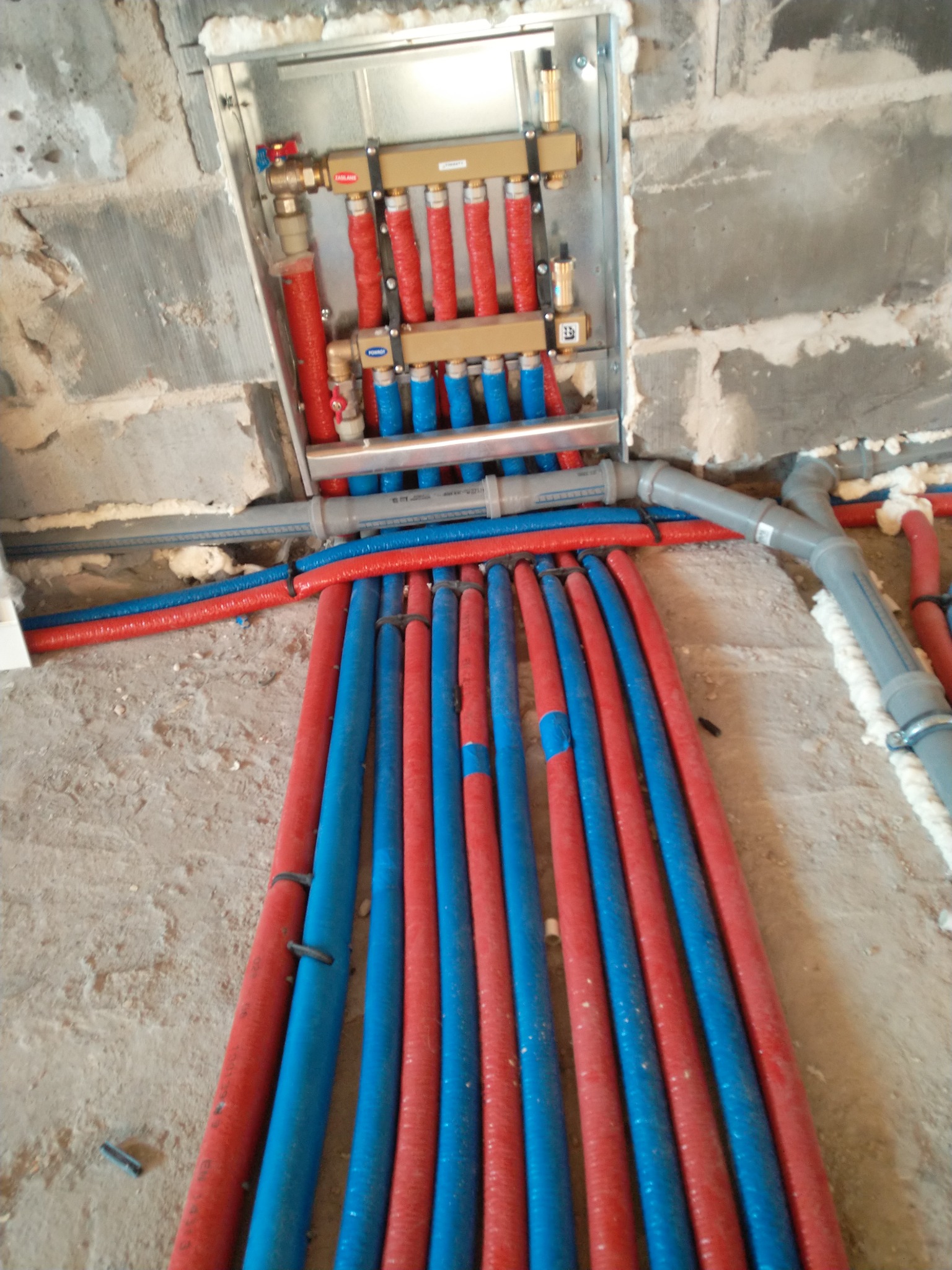 Rozdzielacz instalacji centralnego ogrzewania z rurami w kolorach czerwonym i niebieskim, zamocowany w ścianie z surowej cegły, widoczne elementy mocujące i izolacja piankowa.