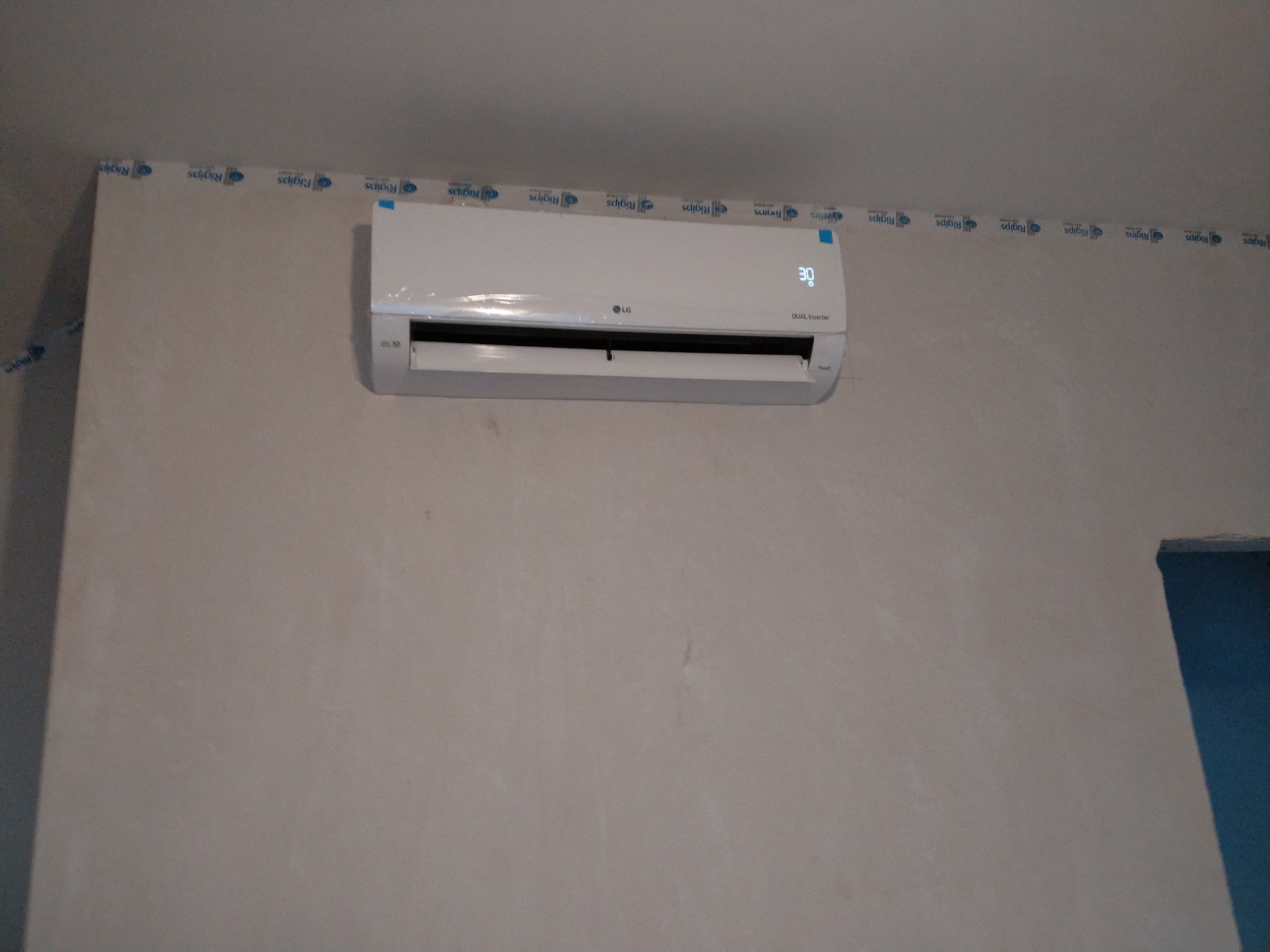 Biały klimatyzator LG Dual Inverter zamontowany na ścianie, z widocznym wyświetlaczem temperatury ustawionym na 30 stopni Celsjusza. Ściana przygotowana do malowania, zabezpieczona taśmą malarską...