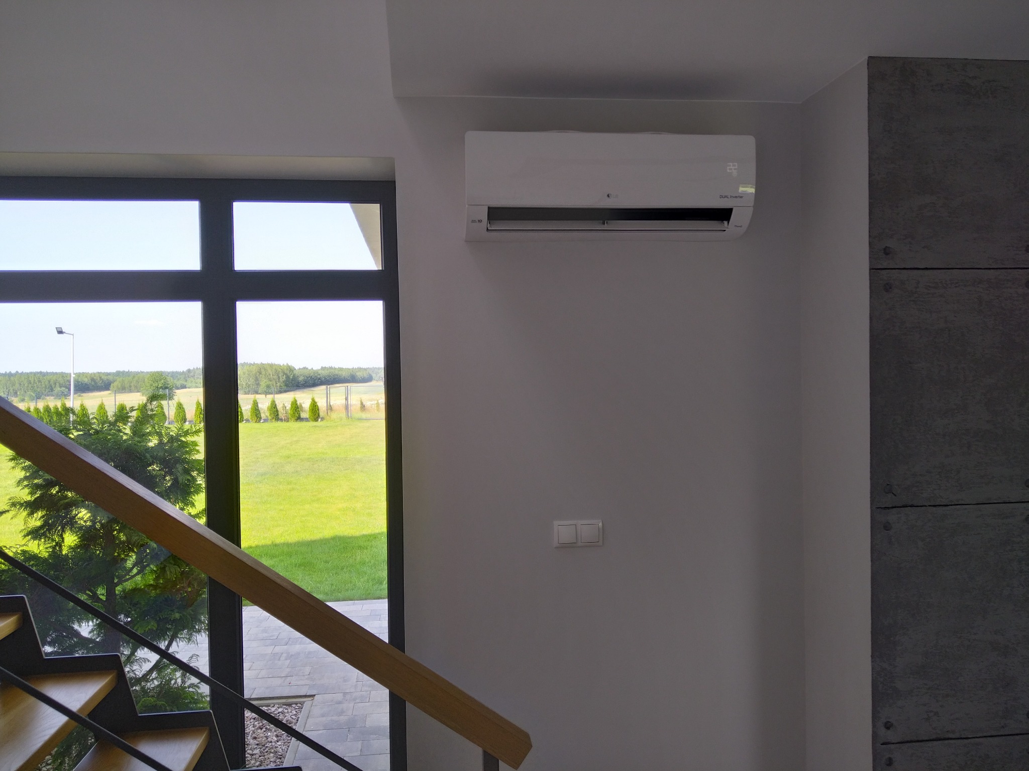 Biały klimatyzator LG Dual Inverter zamontowany na szarej ścianie w nowoczesnym wnętrzu, widok z klatki schodowej na ogród przez duże okno.