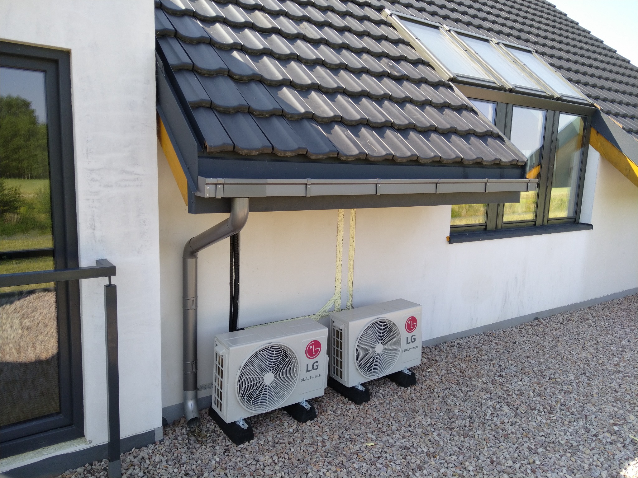 Dwa zewnętrzne moduły klimatyzacji LG Dual Inverter zainstalowane na czarnych podstawach przy ścianie budynku z szarą dachówką i oknami, widoczne rury spustowe i instalacja elektryczna.