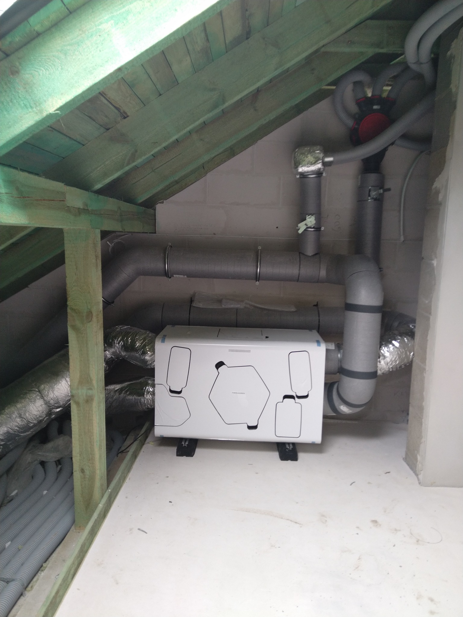 Biała centrala wentylacyjna z rekuperacją, zamontowana na poddaszu, widoczne szare rury wentylacyjne, drewniana konstrukcja dachu i sufit z elementami wykończenia.