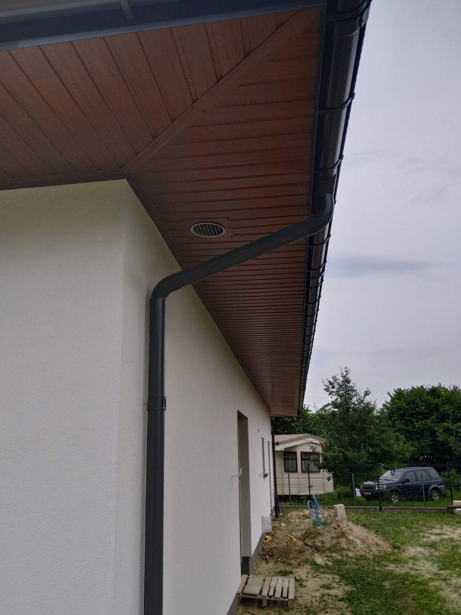 Zewnętrzny róg budynku z zamontowaną, grafitową rynną spustową, widoczna brązowa podbitka dachowa i fragment elewacji. W tle trawnik, drzewa i zaparkowany samochód.