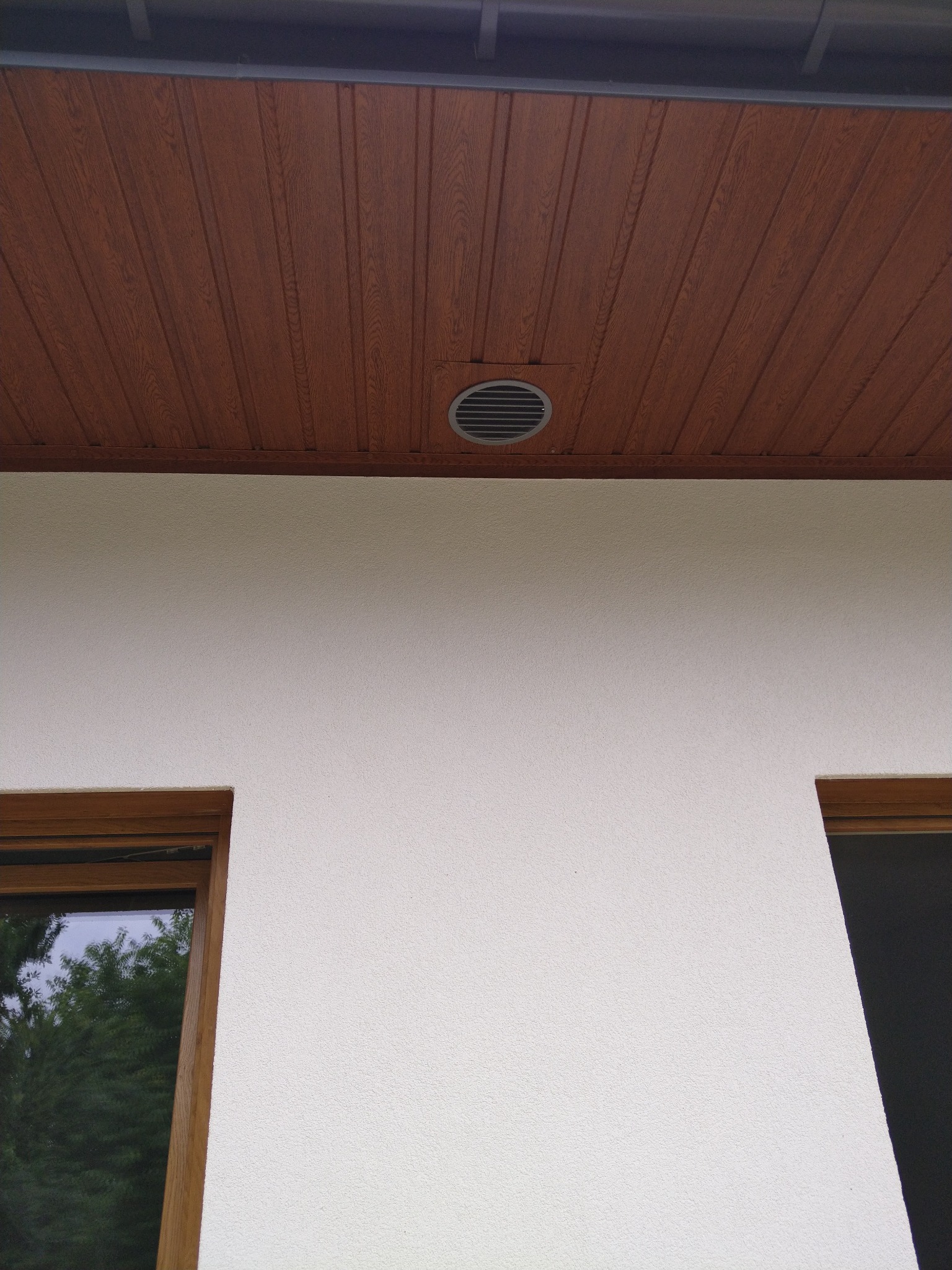 Elewacja budynku z widocznym otworem wentylacyjnym pod drewnianą podbitką dachową, obok fragmenty okien.