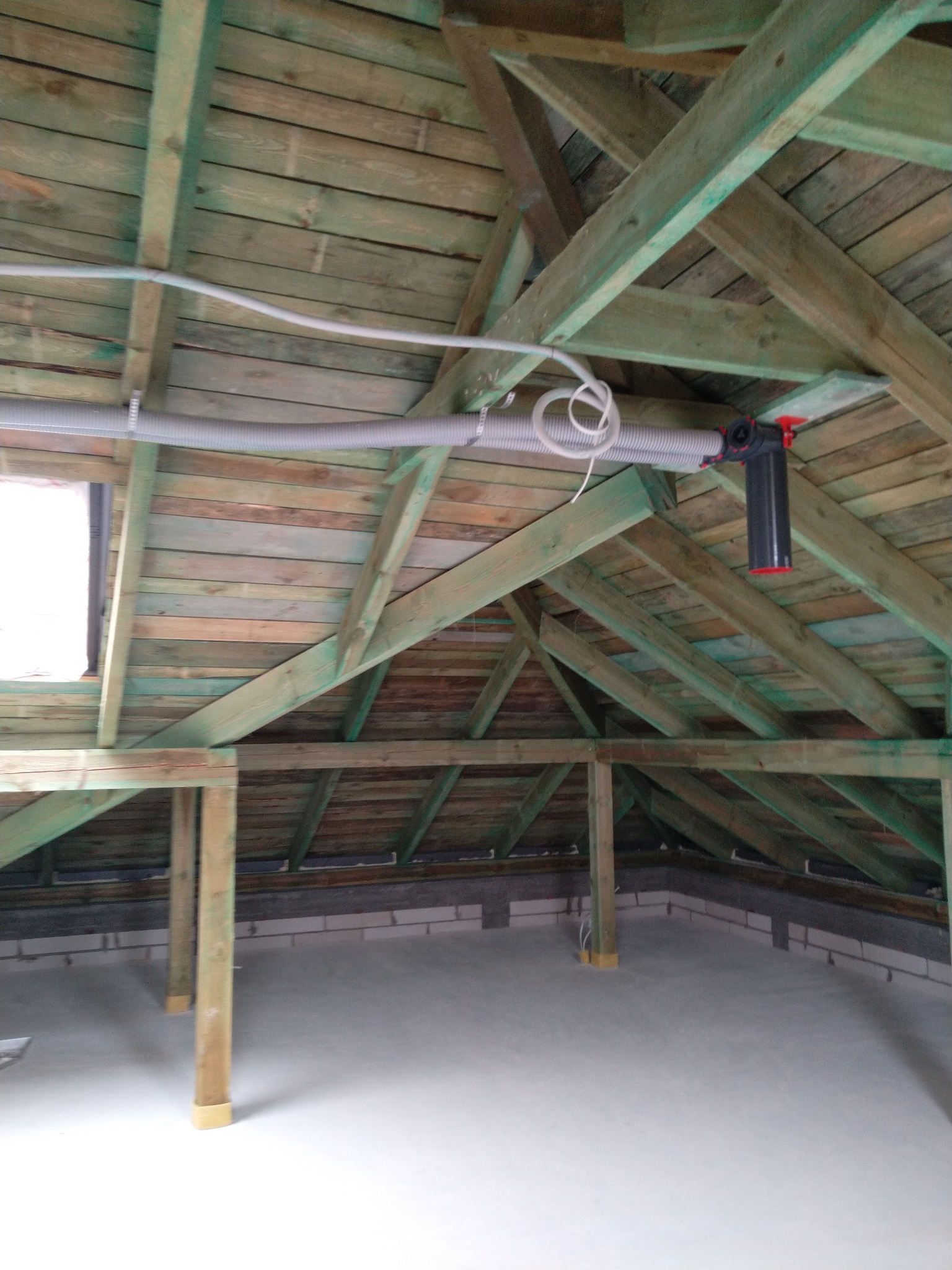 Szare rury wentylacyjne przymocowane do drewnianej konstrukcji dachu na poddaszu, widoczny element montażowy z czerwonym akcentem.