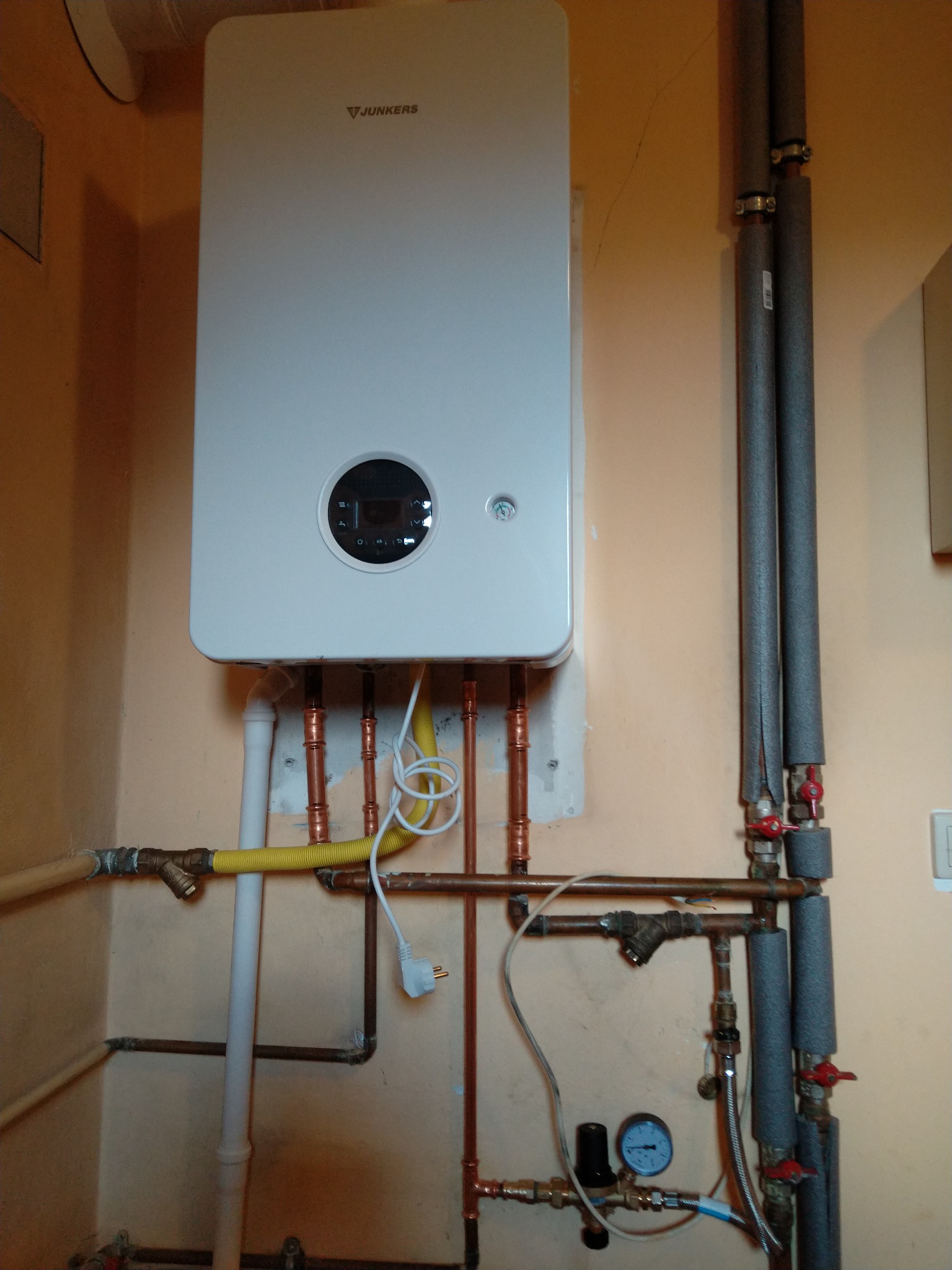 Nowoczesny, biały piec gazowy Junkers zamontowany na ścianie, widoczne liczne rury miedziane, plastikowe i w izolacji, manometr oraz przewód zasilający z wtyczką.