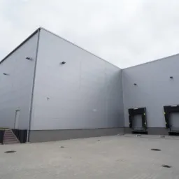 Zewnętrzny widok nowoczesnego magazynu z szarymi panelami, dwoma rampami załadunkowymi z czarnymi odbojnikami i betonowym placem manewrowym.