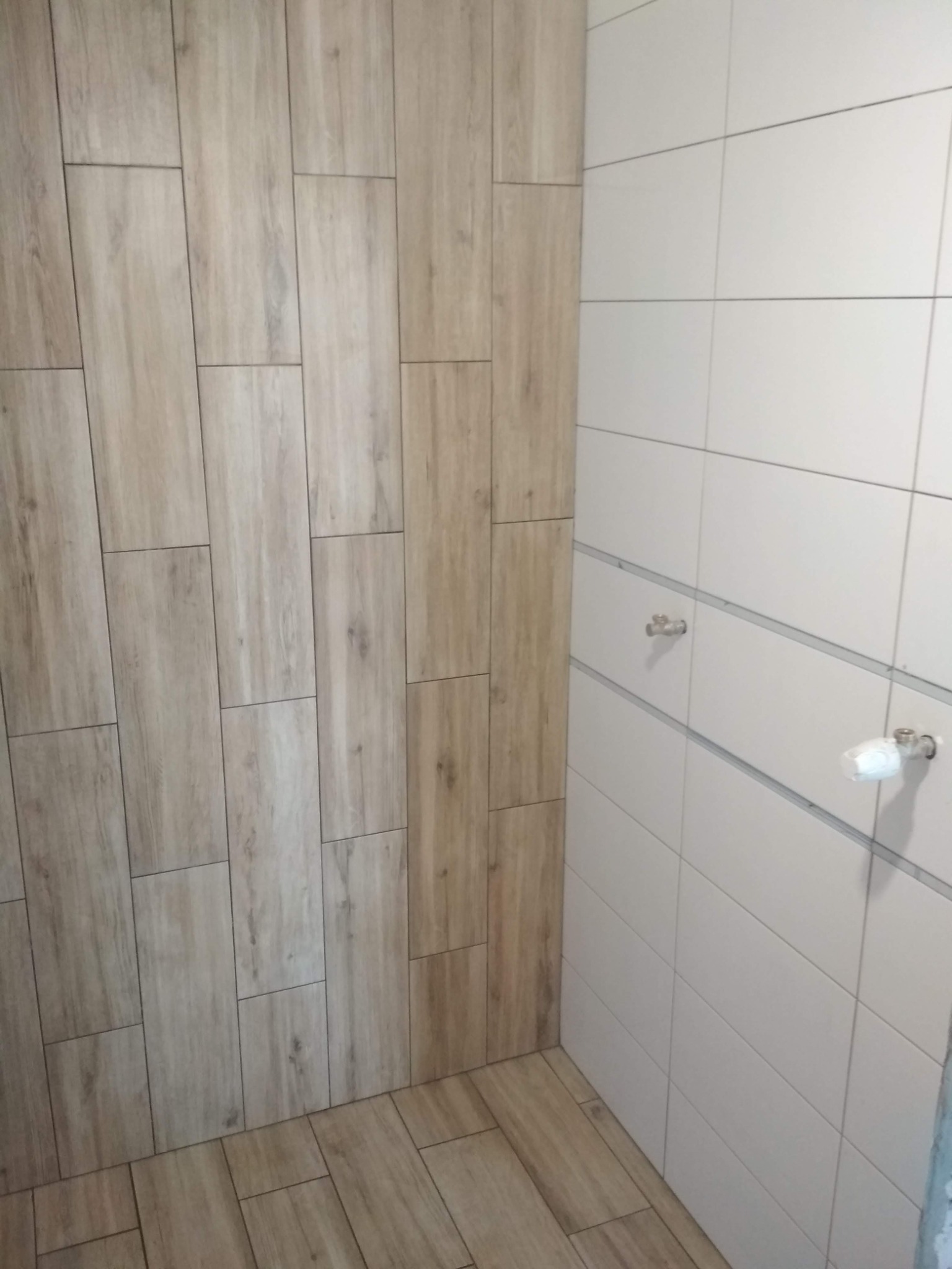 Wykończona ściana i podłoga w łazience z płytek imitujących drewno oraz białych, prostokątnych płytek ściennych z widocznymi zaworami.