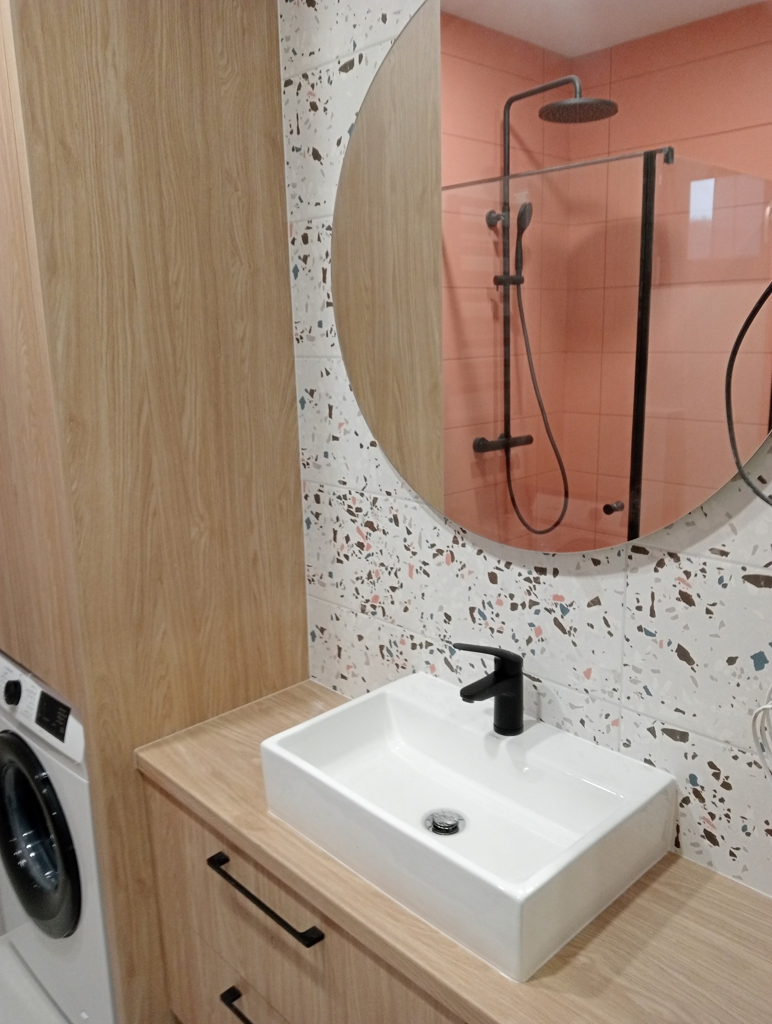 Nowoczesna łazienka z umywalką nablatową, czarną armaturą i płytkami w stylu lastryko na ścianie, odbicie kabiny prysznicowej w okrągłym lustrze.