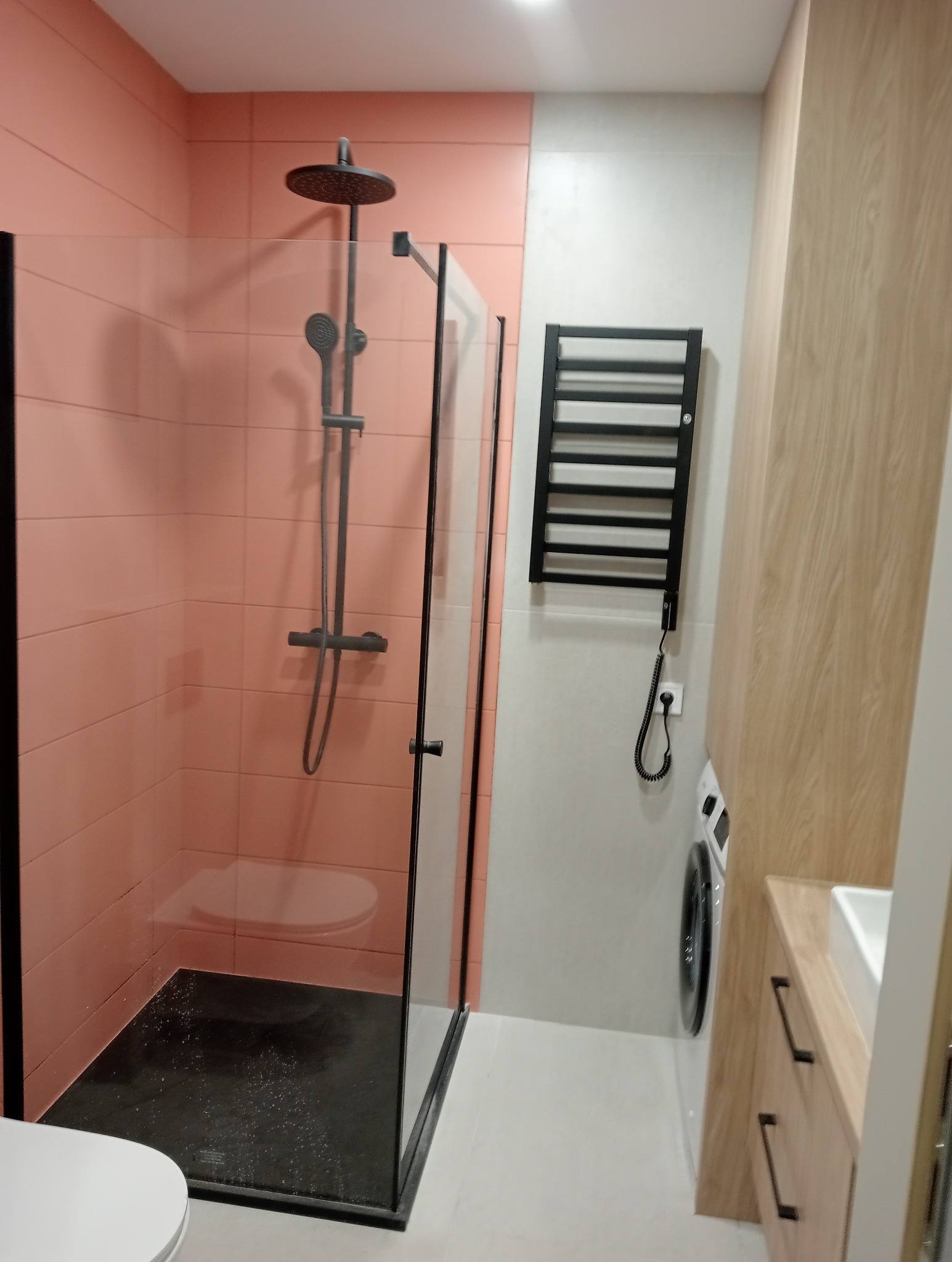 Nowoczesna kabina prysznicowa typu walk-in z czarną armaturą i brodzikiem, różowe płytki na ścianie, grzejnik drabinkowy i pralka w tle.