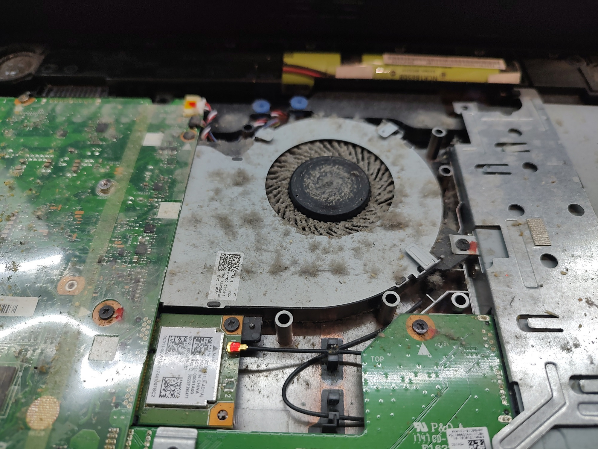 Wnętrze otwartego laptopa z zakurzonym wentylatorem procesora i widoczną płytą główną. Zaznaczone złącze anteny Wi-Fi z czerwonym kablem.