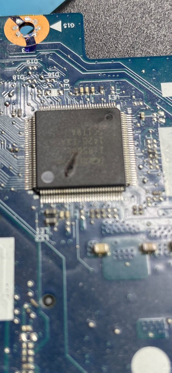 Uszkodzony układ scalony z widocznym przepaleniem na niebieskiej płycie głównej laptopa, widoczne ślady lutowania.