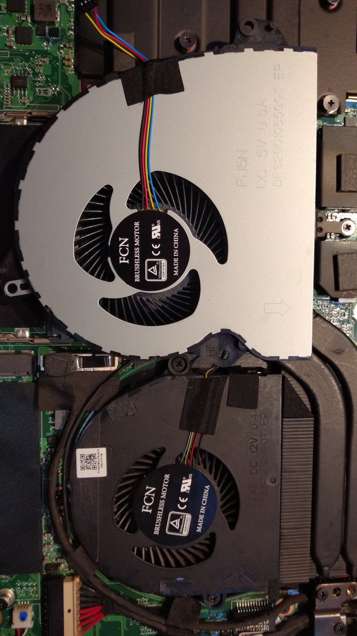 Wnętrze laptopa z widocznymi dwoma wentylatorami chłodzącymi procesor i kartę graficzną, podłączonymi kolorowymi przewodami do płyty głównej. Widoczne oznaczenia producenta FCN i parametry...
