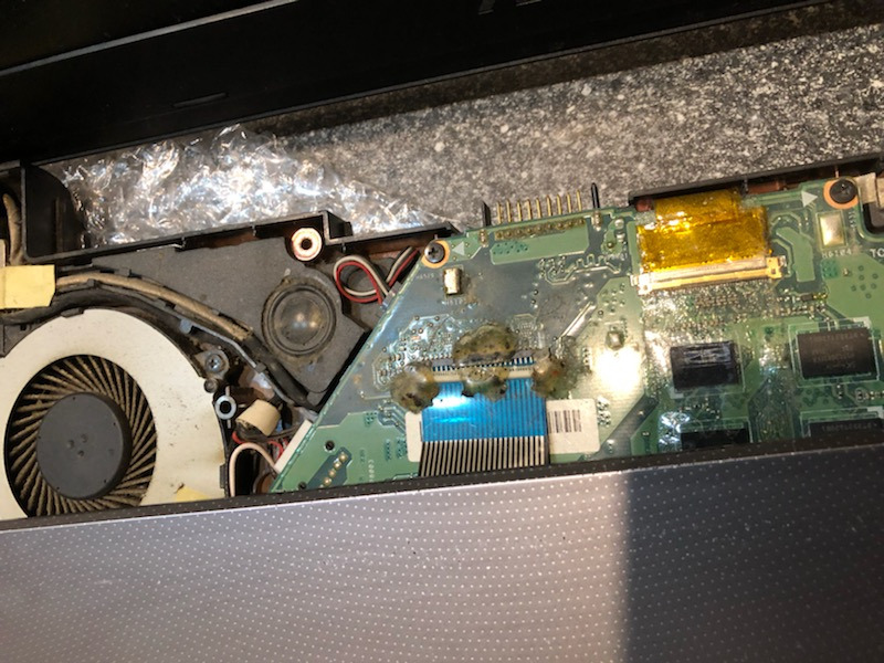 Wnętrze otwartego laptopa z widoczną zakurzoną płytą główną, wentylatorem i elementami chłodzenia, ślady zalania widoczne na płycie.