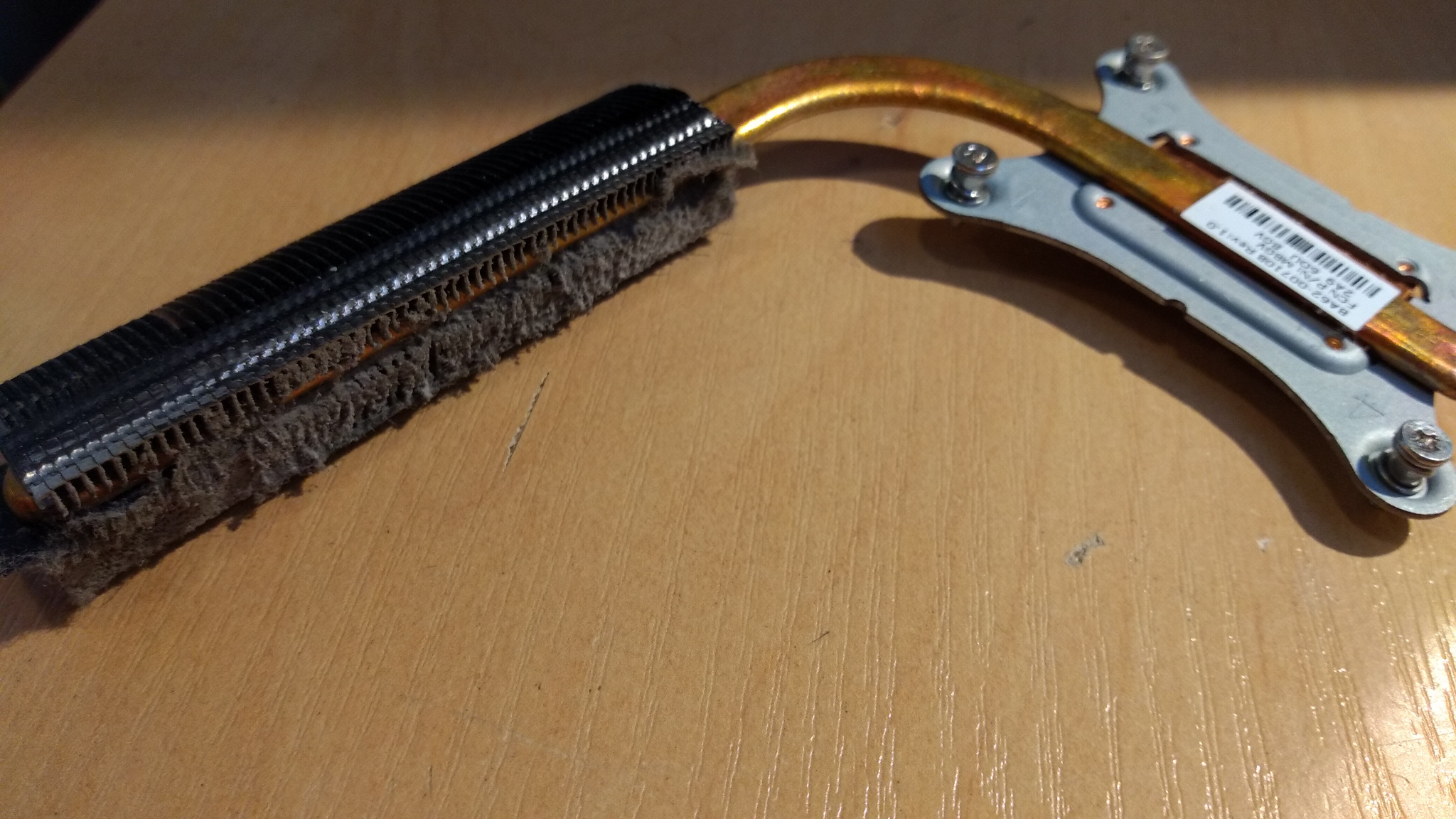 Zabrudzony radiator procesora z widocznymi żeberkami i kurzem, połączony miedzianą rurką cieplną z metalowym mocowaniem z czterema śrubami, leżący na drewnianej powierzchni.