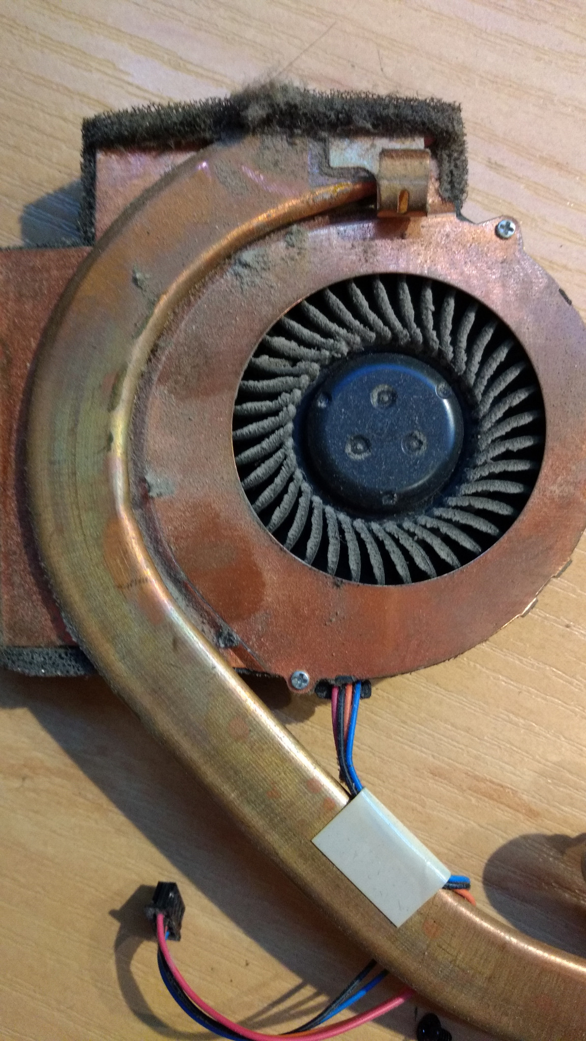Zabrudzony wentylator chłodzenia laptopa z miedzianym radiatorem i widocznymi przewodami, leżący na drewnianym blacie.