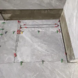 Szare płytki podłogowe z marmurowym wzorem, ułożone z użyciem systemu poziomowania z zielonymi i czerwonymi klipsami, widoczna linia fugi i fragment odpływu liniowego.