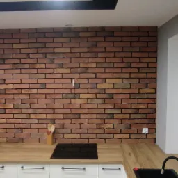 Kuchnia z dekoracyjną ścianą z cegły klinkierowej, drewnianym blatem i białymi szafkami, widoczne okno z białymi ramami i szare zasłony.