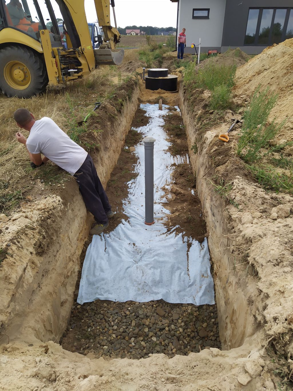 Wykop z ułożoną geowłókniną i warstwą żwiru, widoczna rura pionowa, w tle koparka i zbiorniki na ścieki, pracownik oparty o krawędź wykopu.